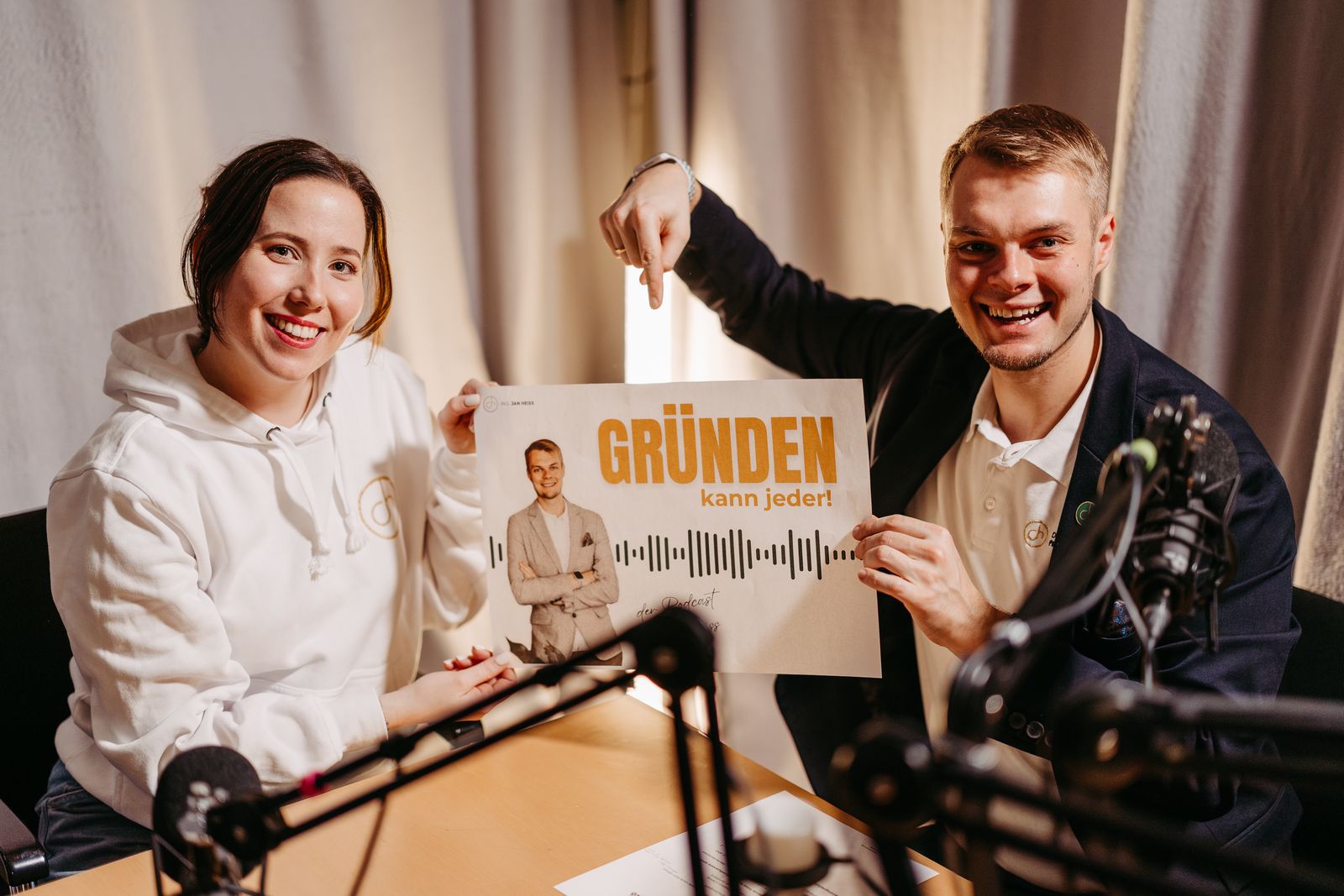 Verena und Jan Heiss lachend im Podcast-Studio mit dem Schild „GRÜNDEN kann jeder!“