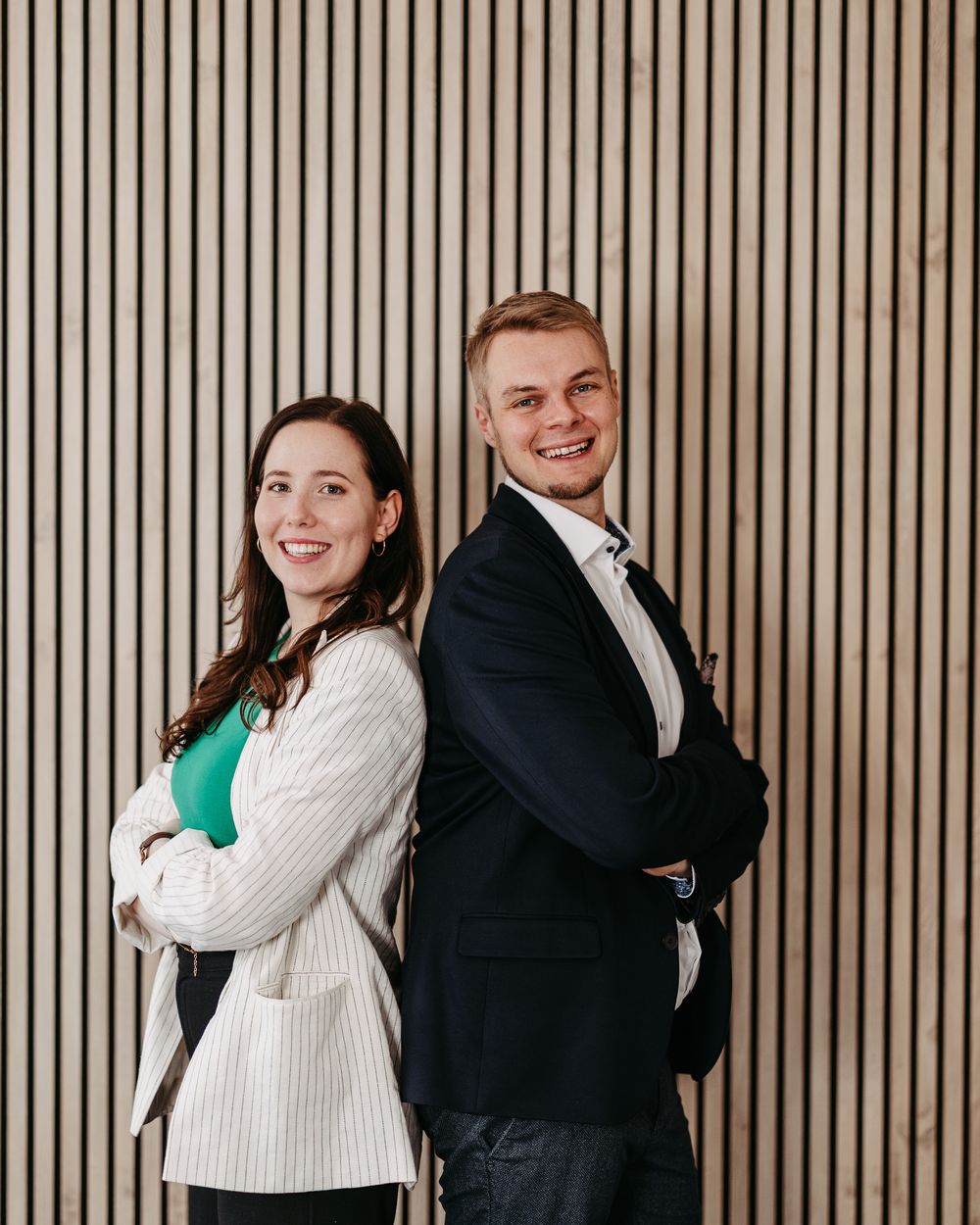 Verena und Jan Heiss stehen Rücken an Rücken vor einer Holzwand und lächeln in die Kamera
